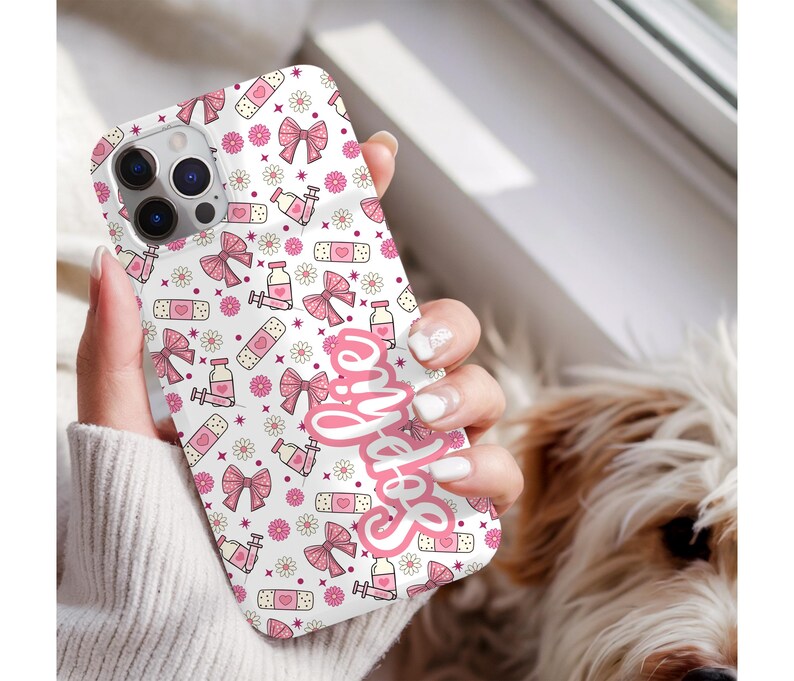 May include: A white phone case with a pink pattern of bows, bandages, flowers, and syringes. The name "Sophie" is written in pink script on the case. The case is held in a hand.