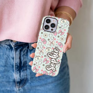 May include: A light blue phone case with a pattern of medical symbols, bows, and flowers in pink and white. The name "Sophie" is written in a cursive font on the case.