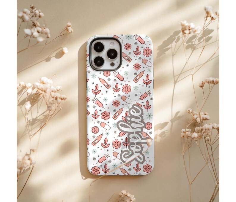 May include: A white phone case with a pattern of pink flowers, syringes, pills, and leaves. The name "Sophie" is written in cursive on the side. The case has a camera cutout.