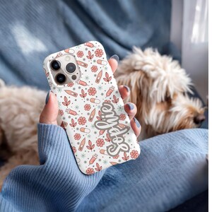 May include: A white phone case with a pattern of pink flowers, pills, syringes, and thermometers. The word "Selfie" is written in a cursive font on the case. The case is held in front of a dog.