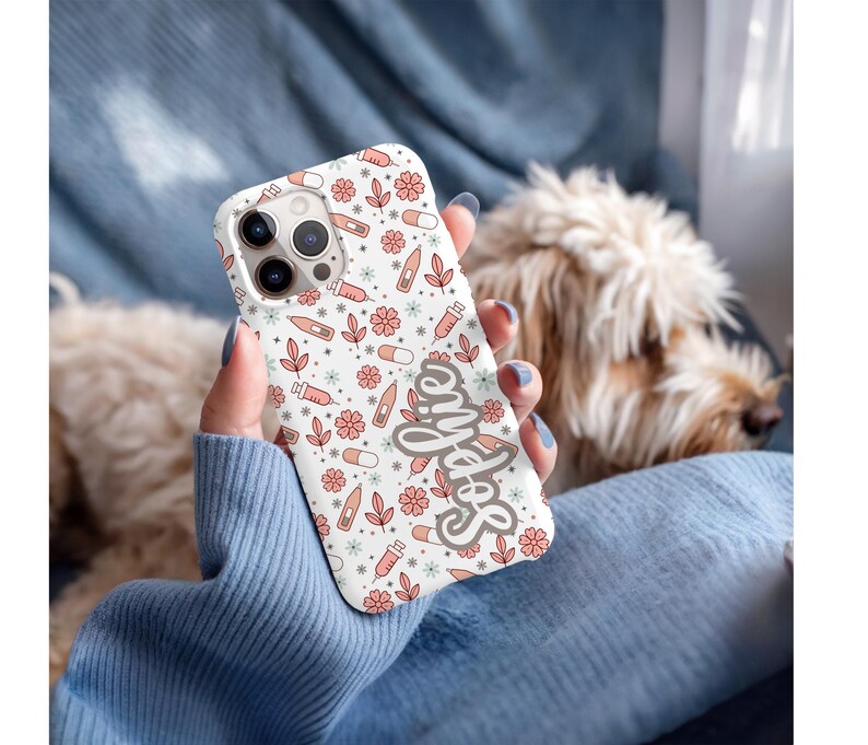 May include: A white phone case with a pattern of pink flowers, pills, syringes, and thermometers. The word "Selfie" is written in a cursive font on the case. The case is held in front of a dog.