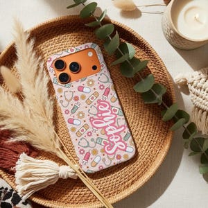 May include: A pink phone case with a pattern of stethoscopes, pills, bandages, and flowers. The name "Sophie" is written in a cursive font across the case. The phone case is on a woven tray with decorative elements.