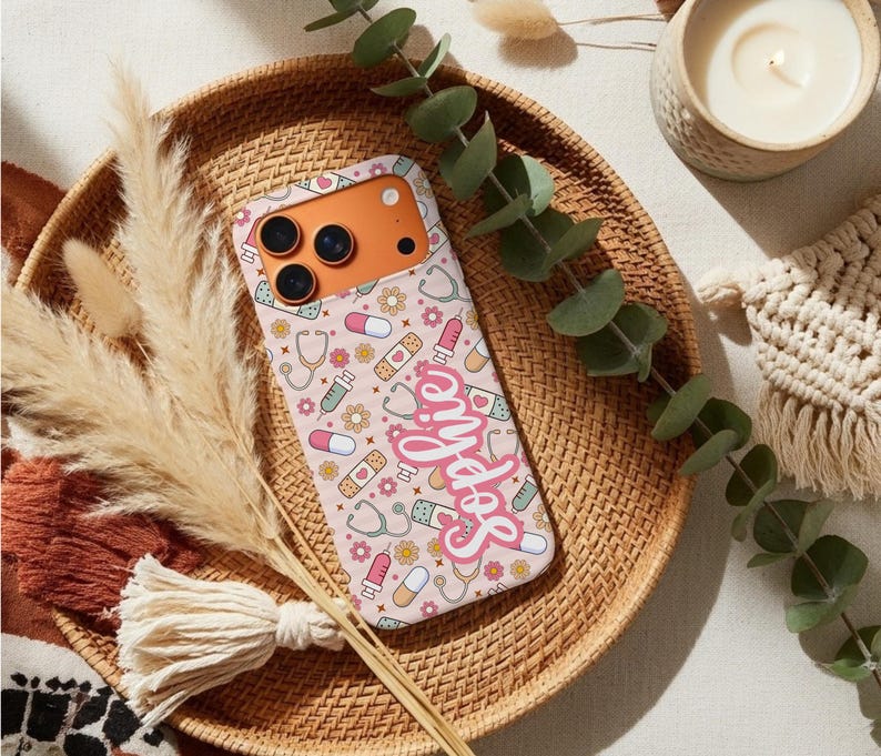 May include: A pink phone case with a pattern of stethoscopes, pills, bandages, and flowers. The name "Sophie" is written in a cursive font across the case. The phone case is on a woven tray with decorative elements.