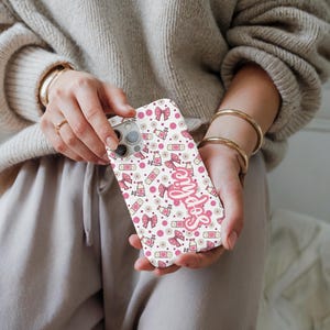 May include: A white phone case with a repeating pattern of pink bows, bottles, and flowers. The word "Selfies" is written in pink script across the case. The case is held in a person's hands.