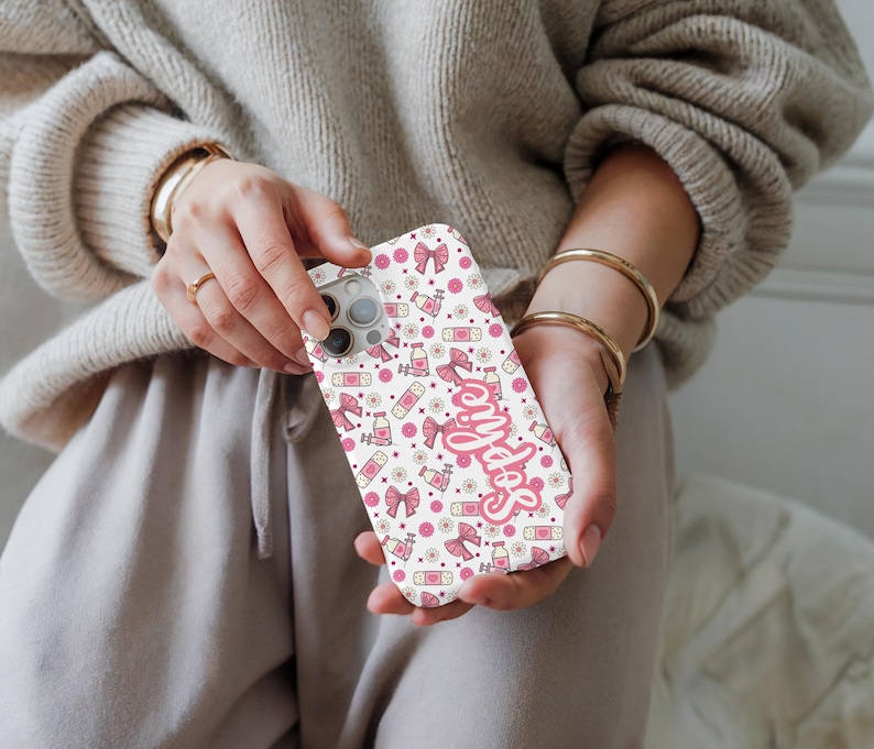 May include: A white phone case with a repeating pattern of pink bows, bottles, and flowers. The word "Selfies" is written in pink script across the case. The case is held in a person's hands.
