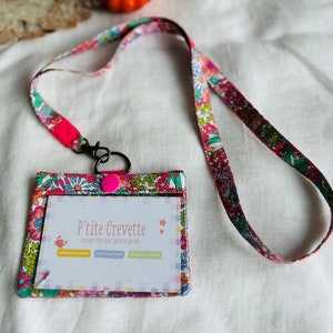 Peut inclure: Un porte-badge avec un cordon floral assorti. Le porte-badge a une fenêtre transparente et une fermeture à bouton rose. Le cordon et le porte-badge présentent un motif floral coloré avec des accents roses, verts et bleus. Le texte "P'tite Crevette" est visible.