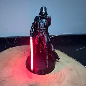 May include: A black Darth Vader figurine with a red lightsaber. The figurine is standing on a round base and is holding the lightsaber in its right hand. The lightsaber glows red. The figurine is on a wooden surface.