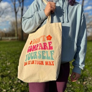 Può includere: Borsa tote color crema con la scritta "DON'T COMPARE YOURSELF DO IT IN YOUR WAY" in stile retrò. La borsa presenta una grafica floreale ed è tenuta da una persona che indossa una felpa con cappuccio blu.