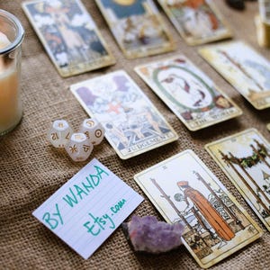 May include: A collection of tarot cards with various illustrations, including The World and Judgement, arranged on a burlap surface. Also present are dice with zodiac symbols, a purple crystal, and a handwritten note that says "BY WANDA etsy.com".