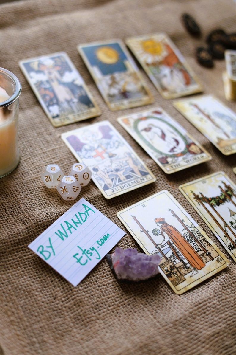 May include: A collection of tarot cards with various illustrations, including The World and Judgement, arranged on a burlap surface. Also present are dice with zodiac symbols, a purple crystal, and a handwritten note that says "BY WANDA etsy.com".