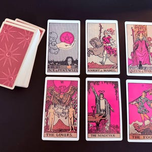 May include: A tarot deck with pink and white cards. The deck includes cards such as "Ace of Pentacles," "Knight of Wands," "Queen of Wands," "The Lovers," "The Magician," and "The Fool." The card backs have a pink design with moons and stars.