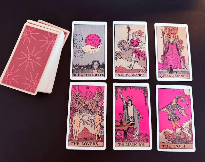 May include: A tarot deck with pink and white cards. The deck includes cards such as "Ace of Pentacles," "Knight of Wands," "Queen of Wands," "The Lovers," "The Magician," and "The Fool." The card backs have a pink design with moons and stars.