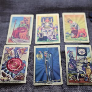 May include: A tarot card reading with six cards laid out, including "Wheel of Fortune" and "The Hermit". The cards have gold borders and colorful illustrations. A lit candle, dice, and crystals are also present, creating a mystical atmosphere.