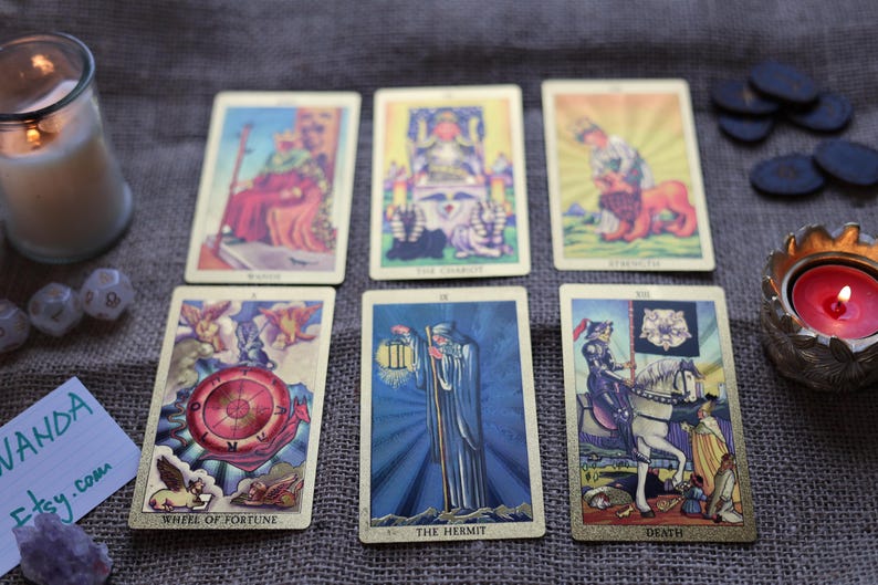 May include: A tarot card reading with six cards laid out, including "Wheel of Fortune" and "The Hermit". The cards have gold borders and colorful illustrations. A lit candle, dice, and crystals are also present, creating a mystical atmosphere.