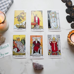 May include: Tarot cards with illustrations and titles such as "The Fool," "The Magician," and "The High Priestess" are displayed. A lit candle and other items are arranged on a white surface, creating a mystical scene.