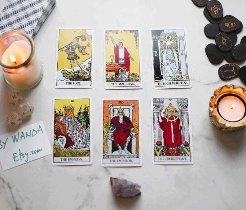 May include: Tarot cards with illustrations and titles such as "The Fool," "The Magician," and "The High Priestess" are displayed. A lit candle and other items are arranged on a white surface, creating a mystical scene.