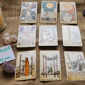 May include: A collection of tarot cards with various illustrations, arranged on a burlap surface. The cards feature images of the sun, moon, and other symbolic figures. Lit candles and crystals are also present, along with dice and a note that says "By Wanda Etsy.com".