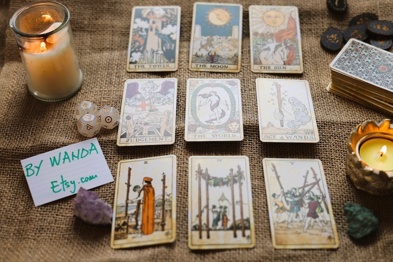 May include: A collection of tarot cards with various illustrations, arranged on a burlap surface. The cards feature images of the sun, moon, and other symbolic figures. Lit candles and crystals are also present, along with dice and a note that says "By Wanda Etsy.com".