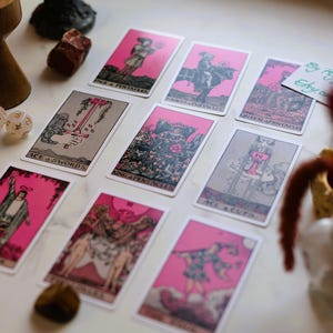 May include: A collection of tarot cards with pink borders and illustrations, arranged on a marble surface. The cards feature various figures and symbols. Other items include a wooden candle holder, dice, and crystals.