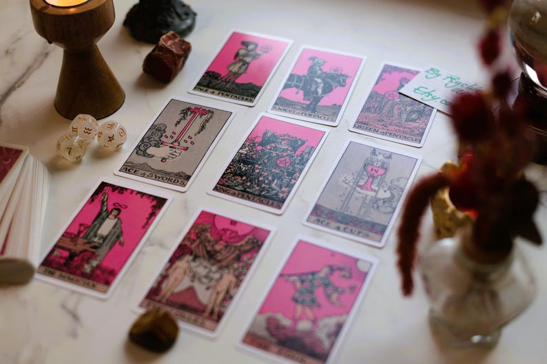 May include: A collection of tarot cards with pink borders and illustrations, arranged on a marble surface. The cards feature various figures and symbols. Other items include a wooden candle holder, dice, and crystals.