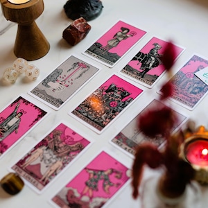 May include: A collection of tarot cards with pink borders and illustrations, arranged on a marble surface. The cards feature various images and titles. Additional items include dice, crystals, and a lit candle in a decorative holder. The text "By Psychic Wanda etsy.com" is visible.