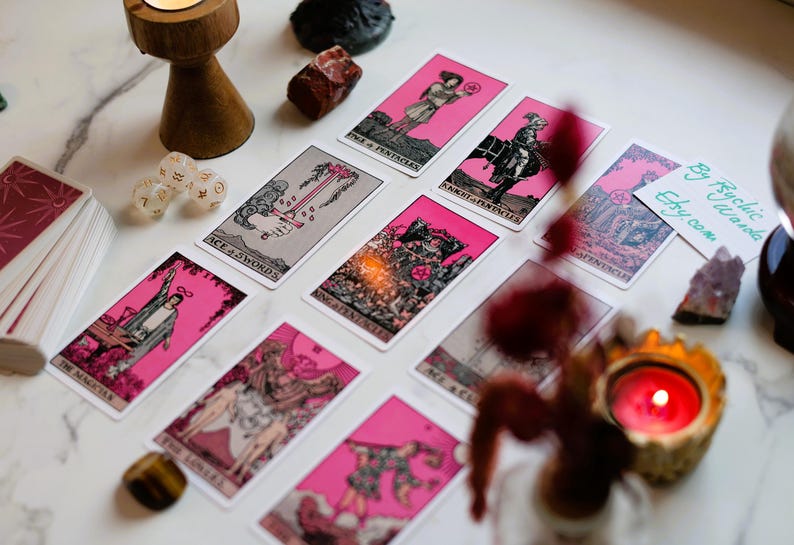 May include: A collection of tarot cards with pink borders and illustrations, arranged on a marble surface. The cards feature various images and titles. Additional items include dice, crystals, and a lit candle in a decorative holder. The text "By Psychic Wanda etsy.com" is visible.