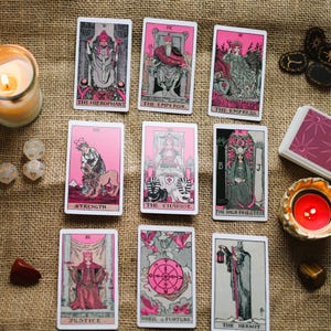 May include: A collection of tarot cards with pink and black illustrations, arranged on a burlap surface. The cards feature images of The Hierophant, The Emperor, The Empress, and others. A lit candle, dice, and crystals are also present.