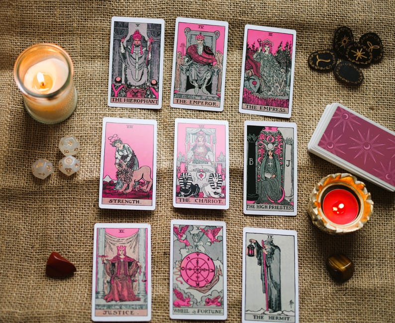 May include: A collection of tarot cards with pink and black illustrations, arranged on a burlap surface. The cards feature images of The Hierophant, The Emperor, The Empress, and others. A lit candle, dice, and crystals are also present.