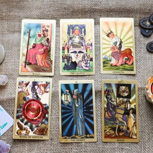 May include: A collection of tarot cards with illustrations, including "The Chariot" and "Strength", arranged on a textured surface. Accompanying items include candles, dice, and decorative stones. A handwritten note reads "By Wanda Etsy.com".