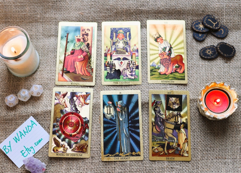 May include: A collection of tarot cards with illustrations, including "The Chariot" and "Strength", arranged on a textured surface. Accompanying items include candles, dice, and decorative stones. A handwritten note reads "By Wanda Etsy.com".