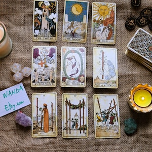 May include: A selection of tarot cards featuring images such as "The Tower," "The Moon," and "The Sun." The cards are displayed alongside a lit candle, dice, crystals, and a deck of cards. The text "BY WANDA Etsy.com" is visible.