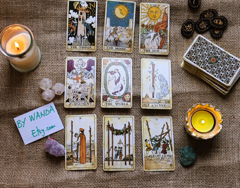 May include: A selection of tarot cards featuring images such as "The Tower," "The Moon," and "The Sun." The cards are displayed alongside a lit candle, dice, crystals, and a deck of cards. The text "BY WANDA Etsy.com" is visible.