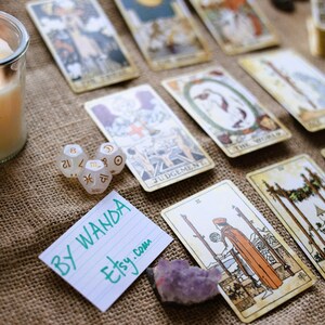 May include: A collection of tarot cards with various illustrations, including The World and Judgement, arranged on a burlap surface. Also present are dice with zodiac symbols, a purple crystal, and a handwritten note that says "BY WANDA etsy.com".