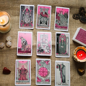 May include: A collection of tarot cards with pink and black illustrations, arranged with candles, dice, and crystals on a burlap surface. The cards feature images of The Hierophant, The Emperor, The Empress, Strength, The Chariot, and others.