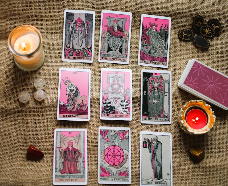 May include: A collection of tarot cards with pink and black illustrations, arranged with candles, dice, and crystals on a burlap surface. The cards feature images of The Hierophant, The Emperor, The Empress, Strength, The Chariot, and others.