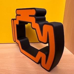 May include: A black and orange decorative engine symbol. The symbol is a three-dimensional object with a black base and orange outline. The object is placed on a light-coloured wooden surface with a yellow background.