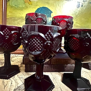 May include: A collection of ruby red goblets with intricate embossed patterns. The goblets have a diamond-like design on the bowl and a detailed stem. The glasses are displayed on a patterned surface, with a painting in the background.