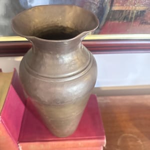May include: A bronze-colored vase with a flared rim and a textured surface. The vase has a rounded body and sits on a stack of red books. The background includes a framed painting.