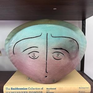 May include: A ceramic sculpture of a face with a teal and pink color scheme. The face has black lines for the eyes, eyebrows, and mouth. The sculpture rests on a book titled "The Smithsonian Collection of Newspaper Comics."