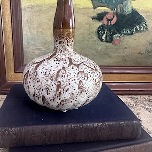 May include: A brown and white speckled ceramic vase with a bulbous base and a long, slender neck. The vase is set on top of two dark blue books. The background features a framed painting with a person holding an umbrella.