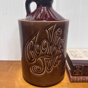 May include: A brown ceramic cookie jar with a handle and a lid. The words "COOKIE JUG" are embossed on the front in a decorative font. The jar is on a wooden surface.