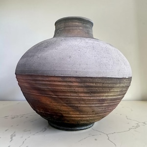 May include: A handcrafted ceramic vase with a bulbous body and a narrow neck. The upper portion is a light gray, while the lower half transitions from brown to reddish-orange. The vase has a textured, ribbed design.