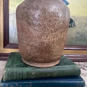 May include: A textured, brown ceramic vase with a wide base and a slightly flared rim. The vase sits atop a stack of three vintage books with green and blue covers. The books have gold lettering.