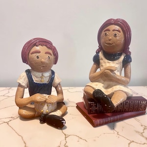 May include: Two ceramic figurines of children. One is seated, knitting, wearing a blue jumper over a white shirt. The other is seated on books, wearing a white dress with blue sleeves. Both have red hair and are painted in earthy tones.