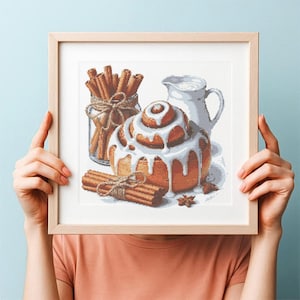 Peut inclure: Une œuvre d'art au point de croix encadrée représentant un pain à la cannelle glacé, un pot de bâtons de cannelle et une cruche blanche. L'œuvre est dans un cadre en bois clair, tenue par une personne portant un t-shirt pêche.