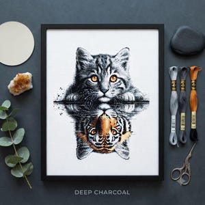 Puede incluir: Un bordado en punto de cruz enmarcado que muestra un gato atigrado gris con ojos color ámbar reflejado en el agua como un tigre con rayas naranjas y negras. La obra se presenta con hilos de bordar en gris, carbón y naranja, además de tijeras.
