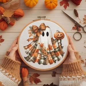 May include: A cross-stitch embroidery hoop featuring a Halloween ghost design. The ghost wears a plaid shawl and holds a carved pumpkin. The design incorporates orange, brown, and white colors, with autumn leaves and a bow. The hoop is held by hands.