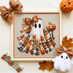 Puede incluir: Un bordado a punto de cruz enmarcado de un fantasma con un chal a cuadros y sosteniendo una calabaza tallada. El fantasma tiene ojos negros y está decorado con hojas de otoño y un lazo. Un fantasma de peluche, lazos y calabazas rodean la obra enmarcada.