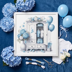 Peut inclure: Un motif de point de croix encadré représentant une armoire blanche remplie de vêtements de bébé, entourée d'hortensias bleus, de ballons et de cadeaux. L'illustration comprend un vase de fleurs bleues, des petits chaussons et des vêtements pliés.