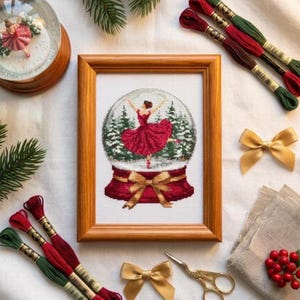 Op de afbeelding: Een ingelijste kruissteek van een ballerina in een sneeuwbol, gekleed in een rode jurk, met een gouden strik. De afbeelding is omgeven door rood en groen borduurgaren, gouden strikken en een kleine gouden schaar. Een sneeuwbol staat in de linkerbovenhoek.